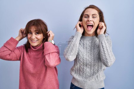 Photo for Mother and daughter standing over blue background smiling pulling ears with fingers, funny gesture. audition problem - Royalty Free Image