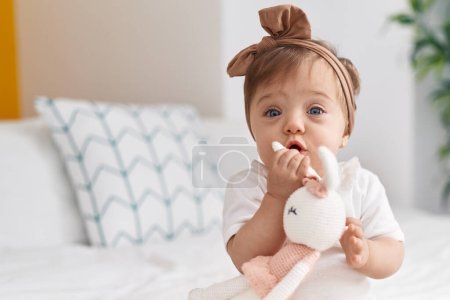 Adorable caucasian baby sitting on bed bitting doll at bedroom