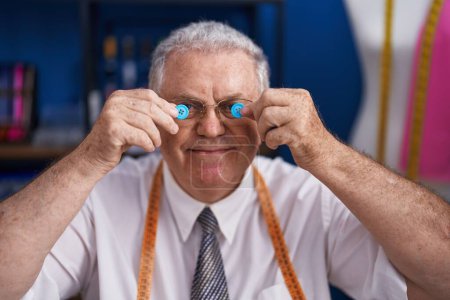 Foto de Hombre de pelo gris de mediana edad sastre sonriendo confiado sosteniendo botones sobre los ojos en la sastrería - Imagen libre de derechos