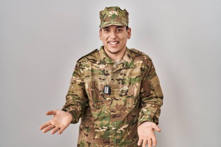 Téléchargez les photos : Jeune homme arabe portant uniforme de camouflage de l'armée souriant gai avec les bras ouverts comme accueil amical, salutations positives et confiantes - en image libre de droit