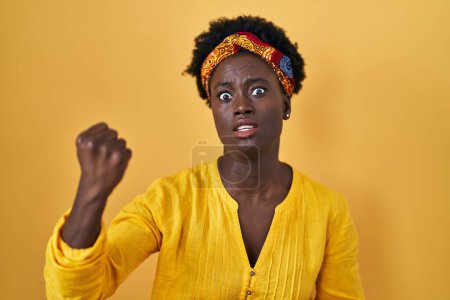 Foto de Mujer joven africana vistiendo turbante africano enojado y loco levantando el puño frustrado y furioso mientras gritaba con ira. rabia y concepto agresivo. - Imagen libre de derechos