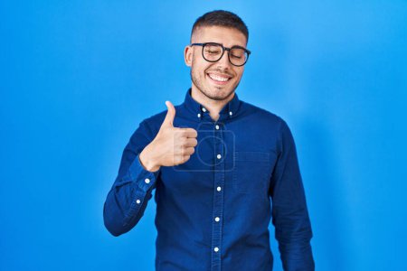 Téléchargez les photos : Jeune homme hispanique portant des lunettes sur fond bleu faisant des pouces heureux geste avec la main. approuvant l'expression regardant la caméra montrant le succès. - en image libre de droit