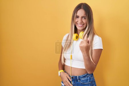 Téléchargez les photos : Jeune femme blonde debout sur fond jaune portant des écouteurs signe venir ici geste avec la main invitant accueillant heureux et souriant - en image libre de droit