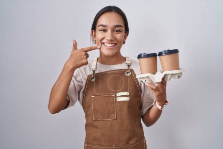 Téléchargez les photos : Jeune femme hispanique portant tablier de serveuse professionnelle tenant café souriant joyeuse montrant et pointant avec les doigts les dents et la bouche. concept de santé dentaire. - en image libre de droit