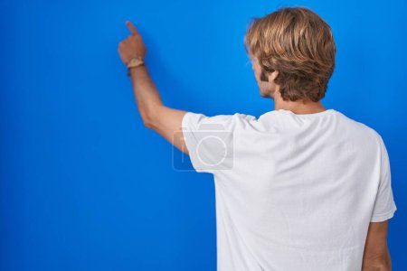 Foto de Hombre de mediana edad de pie sobre fondo azul posando hacia atrás apuntando hacia adelante con la mano del dedo - Imagen libre de derechos