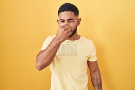 Téléchargez les photos : Jeune homme hispanique debout sur fond jaune sentant quelque chose de puant et dégoûtant, odeur intolérable, retenant souffle avec les doigts sur le nez. mauvaise odeur - en image libre de droit