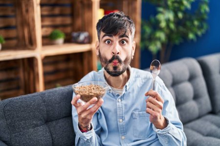 Foto de Joven hombre hispano con barba comiendo cereales integrales saludables haciendo cara de pez con boca y ojos entrecerrados, loco y cómico. - Imagen libre de derechos