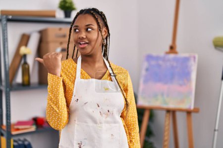 Téléchargez les photos : Femme afro-américaine avec des tresses au studio d'art souriant avec le visage heureux regardant et pointant vers le côté avec le pouce vers le haut. - en image libre de droit