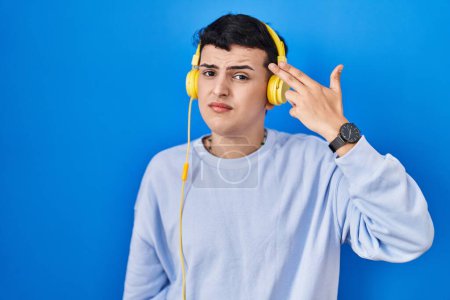 Photo for Non binary person listening to music using headphones shooting and killing oneself pointing hand and fingers to head like gun, suicide gesture. - Royalty Free Image