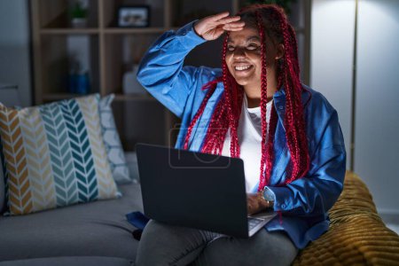 Photo for African american woman with braided hair using computer laptop at night very happy and smiling looking far away with hand over head. searching concept. - Royalty Free Image