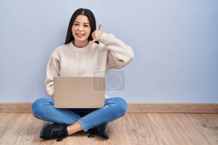 Foto de Mujer joven usando el portátil sentado en el suelo en casa sonriendo haciendo el gesto del teléfono con la mano y los dedos como hablar por teléfono. comunicar conceptos. - Imagen libre de derechos