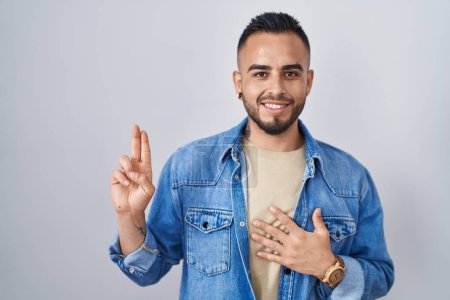 Téléchargez les photos : Jeune homme hispanique debout sur fond isolé souriant jurer avec la main sur la poitrine et les doigts vers le haut, faisant un serment de fidélité promesse - en image libre de droit