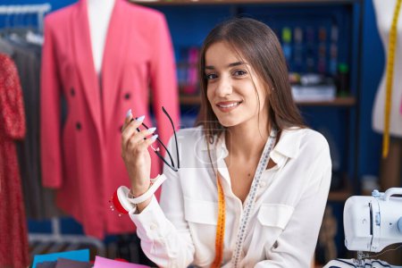 Photo for Young beautiful hispanic woman tailor smiling confident sitting on table at clothing factory - Royalty Free Image