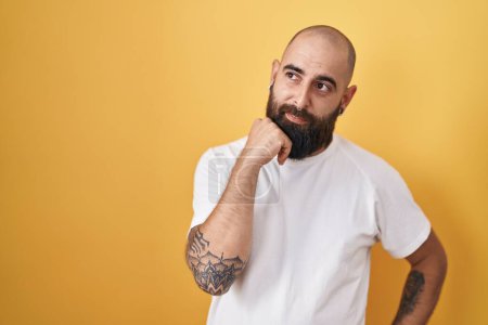 Photo for Young hispanic man with beard and tattoos standing over yellow background with hand on chin thinking about question, pensive expression. smiling with thoughtful face. doubt concept. - Royalty Free Image