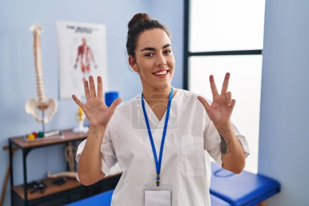 Young hispanic woman working at rehabilitation clinic showing and pointing up with fingers number eight while smiling confident and happy.