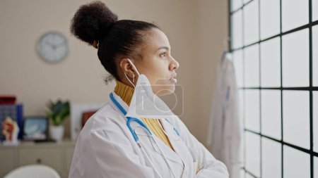 Photo for Young african american woman doctor looking at the window wearing mask at clinic - Royalty Free Image