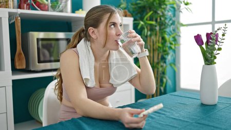 Photo for Young blonde woman wearing sportswear drinking glass of water eating rice cake at dinning room - Royalty Free Image