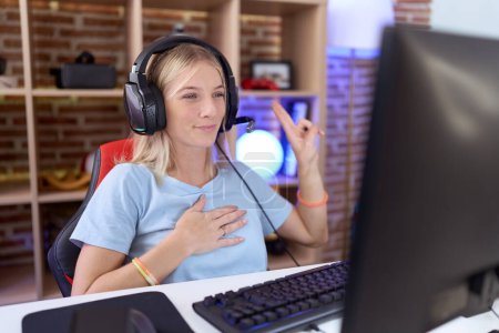Téléchargez les photos : Jeune femme caucasienne jouant à des jeux vidéo portant des écouteurs souriant jurant avec la main sur la poitrine et les doigts vers le haut, faisant un serment de fidélité - en image libre de droit