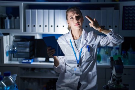 Photo for Beautiful blonde woman working at scientist laboratory late at night shooting and killing oneself pointing hand and fingers to head like gun, suicide gesture. - Royalty Free Image