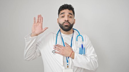 Téléchargez les photos : Jeune homme hispanique médecin faire un serment avec la main sur la poitrine sur fond blanc isolé - en image libre de droit