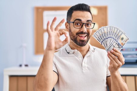 Téléchargez les photos : Jeune homme hispanique avec barbe et lunettes tenant billets en dollars faisant signe ok avec les doigts, sourire geste amical excellent symbole - en image libre de droit