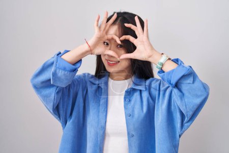 Photo for Young chinese woman standing over white background doing heart shape with hand and fingers smiling looking through sign - Royalty Free Image