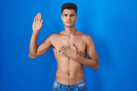 Foto de Joven hombre hispano de pie sin camisa sobre fondo azul jurando con la mano en el pecho y la palma abierta, haciendo un juramento de lealtad promesa - Imagen libre de derechos