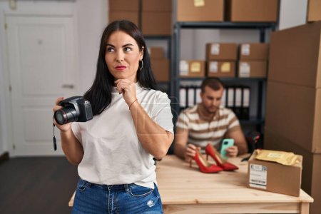 Photo for Young hispanic woman holding camera working at small business ecommerce serious face thinking about question with hand on chin, thoughtful about confusing idea - Royalty Free Image
