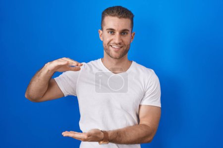 Téléchargez les photos : Jeune homme caucasien debout sur fond bleu geste avec les mains montrant grand et grand signe de taille, symbole de mesure. souriant en regardant la caméra. concept de mesure. - en image libre de droit