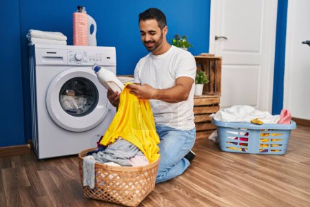 Photo for Young hispanic man washing clothes holding detergent bottle at laundry room - Royalty Free Image