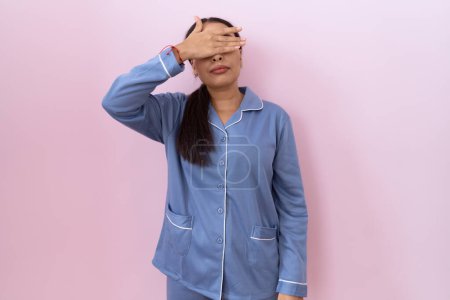 Téléchargez les photos : Jeune femme arabe portant un pyjama bleu couvrant les yeux avec la main, l'air grave et triste. concept de non-visibilité, de dissimulation et de rejet - en image libre de droit