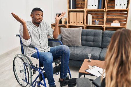 Photo for African american man doing therapy sitting on wheelchair clueless and confused expression with arms and hands raised. doubt concept. - Royalty Free Image