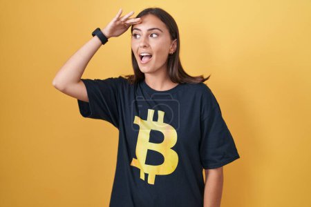 Photo for Young hispanic woman wearing bitcoin t shirt very happy and smiling looking far away with hand over head. searching concept. - Royalty Free Image