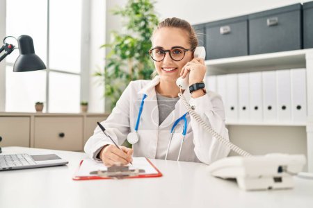 Téléchargez les photos : Jeune femme blonde portant un uniforme de médecin parlant au téléphone travaillant à la clinique - en image libre de droit