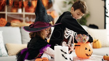 Foto de Adorable chico y chica teniendo fiesta de halloween poniendo caramelos en cesta de calabaza en casa - Imagen libre de derechos
