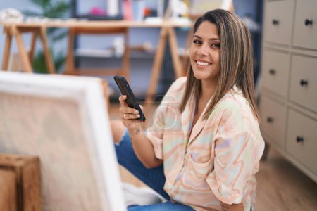Photo for Young hispanic woman artist using smartphone sitting on floor at art studio - Royalty Free Image