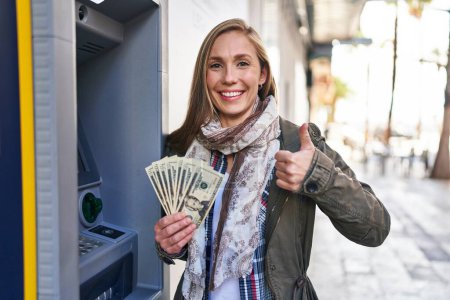 Photo for Young blonde woman holding dollars banknotes from atm machine smiling happy and positive, thumb up doing excellent and approval sign - Royalty Free Image