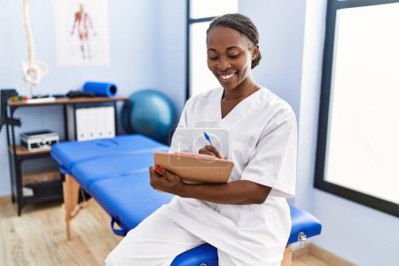 Photo for African american woman physiotherapist smiling confident writing medical report at rehab clinic - Royalty Free Image