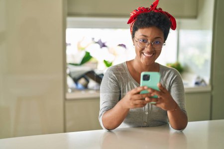 Téléchargez les photos : Femme afro-américaine utilisant smartphone assis sur la table à la maison - en image libre de droit
