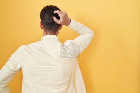 Foto de Young hispanic man standing over yellow background backwards thinking about doubt with hand on head - Imagen libre de derechos