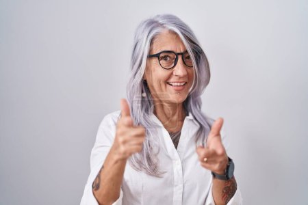 Foto de Mujer de mediana edad con tatuajes con gafas de pie sobre fondo blanco señalando los dedos a la cámara con la cara feliz y divertida. buena energía y vibraciones. - Imagen libre de derechos