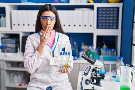 Foto de Young brunette woman working at scientist laboratory covering mouth with hand, shocked and afraid for mistake. surprised expression - Imagen libre de derechos