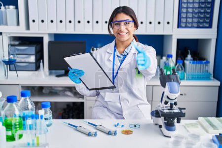 Foto de Mujer joven hispana trabajando en laboratorio científico sonriendo amistosa ofreciendo apretón de manos como saludo y bienvenida. negocios exitosos. - Imagen libre de derechos