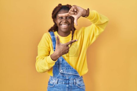 Foto de African woman standing over yellow background smiling making frame with hands and fingers with happy face. creativity and photography concept. - Imagen libre de derechos