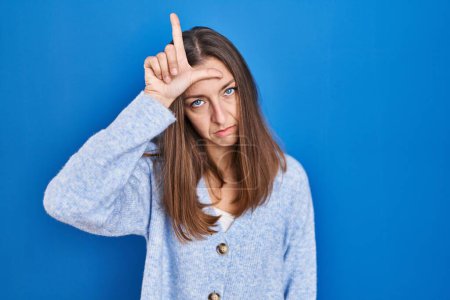Photo for Young woman standing over blue background making fun of people with fingers on forehead doing loser gesture mocking and insulting. - Royalty Free Image