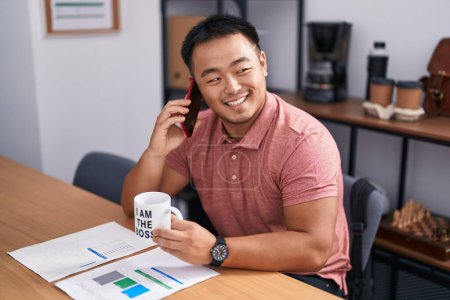 Foto de Young chinese man business worker talking on smartphone drinking coffee at office - Imagen libre de derechos