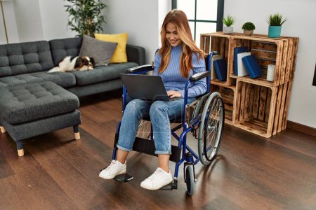 Téléchargez les photos : Jeune femme caucasienne utilisant un ordinateur portable assis sur un fauteuil roulant à la maison - en image libre de droit