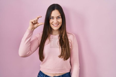 Téléchargez les photos : Young brunette woman standing over pink background smiling and confident gesturing with hand doing small size sign with fingers looking and the camera. measure concept. - en image libre de droit