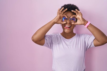 Foto de Joven mujer afroamericana de pie sobre fondo rosa haciendo buen gesto como prismáticos sacando la lengua, ojos mirando a través de los dedos. expresión loca. - Imagen libre de derechos