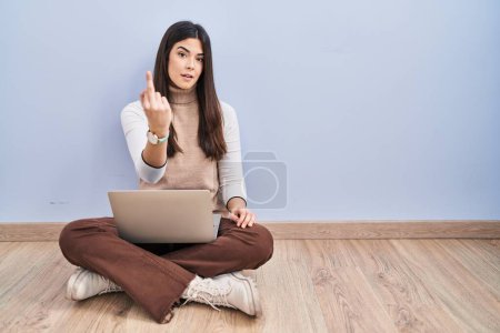 Photo for Young brunette woman working using computer laptop sitting on the floor showing middle finger, impolite and rude fuck off expression - Royalty Free Image